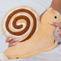 A plush snail pillow being held by hands against a white fabric background. The snail has a soft beige body and a spiral shell pattern in brown and cream.