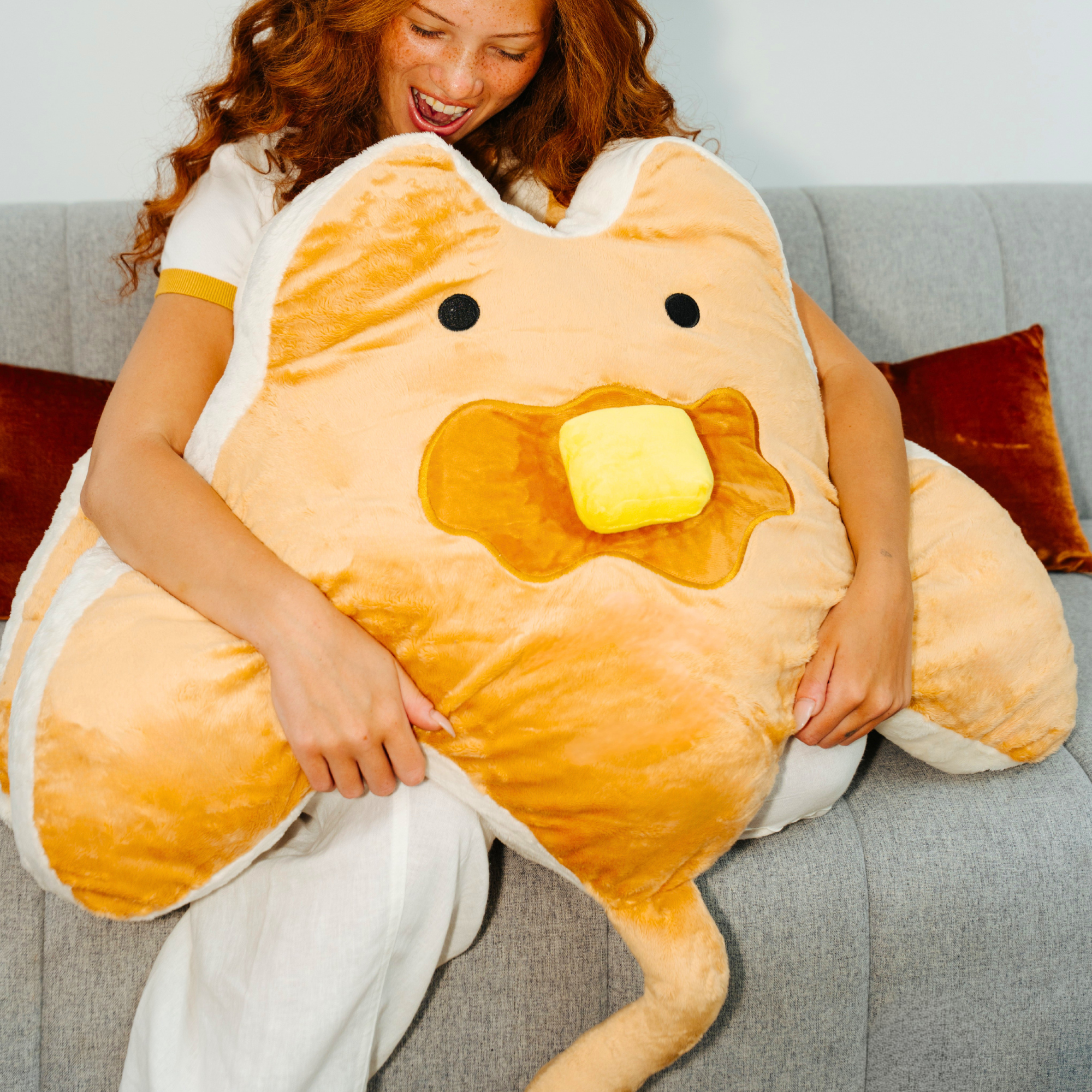 A woman with red hair sits on a gray couch hugging a large pancake manta ray plush shaped pillow. The pillow is made of soft tan and orange fabric with white edges, black embroidered eyes, and a yellow slab of butter.